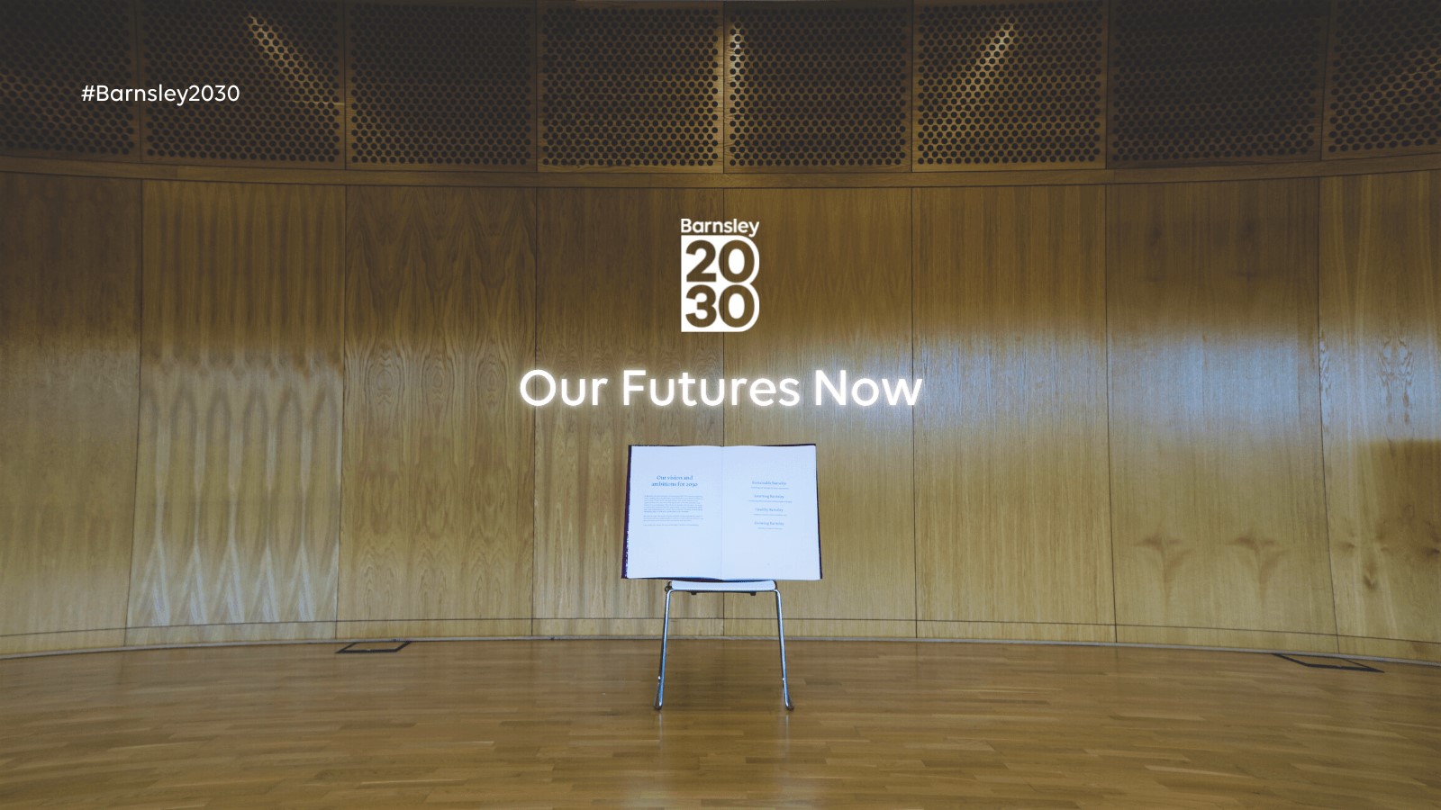 'Our Futures Now' in white neon light above an easel with a large, open book on it
