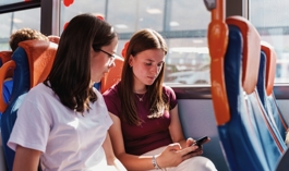 Two Girls On A Bus Looking Down At A Mobile Phone