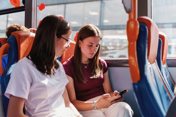 Two Girls On A Bus Looking Down At A Mobile Phone