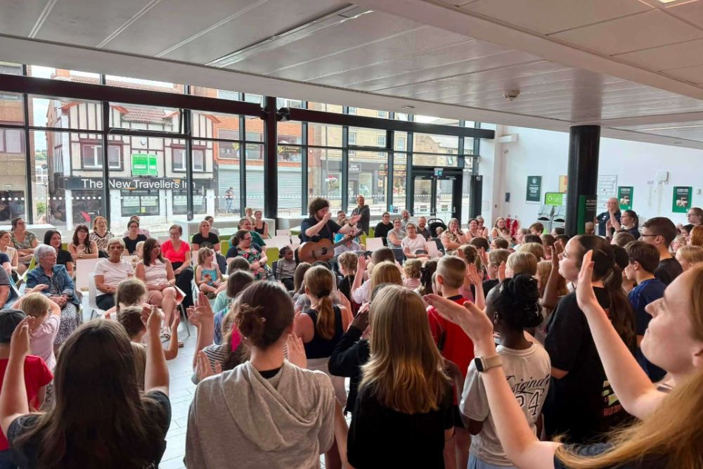 Crowd Of People Standing In A Covered Hall Facing A Seated Group, With Hands Raised As If Cheering Or Responding, Large Windows And Green Posters In The Background