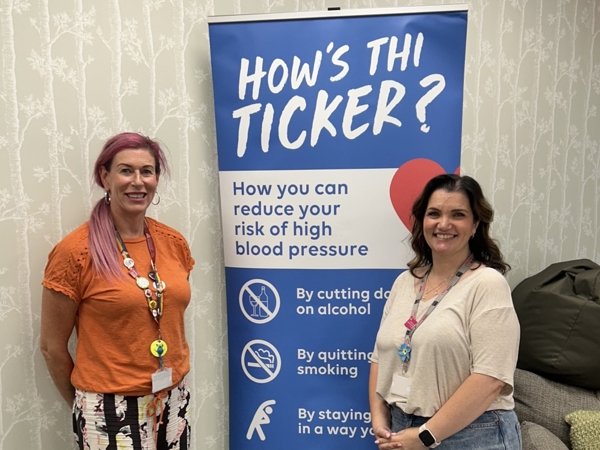 Two women standing by a banner reading How's Thi Ticker.jpg