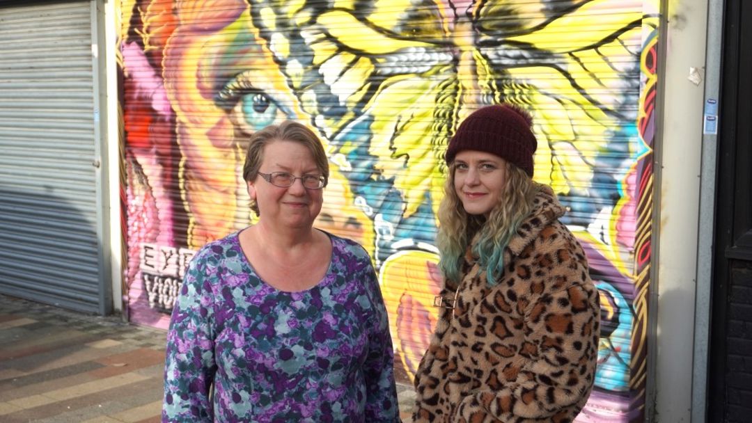 Two women standing in front of a shutter painted with a butterfly